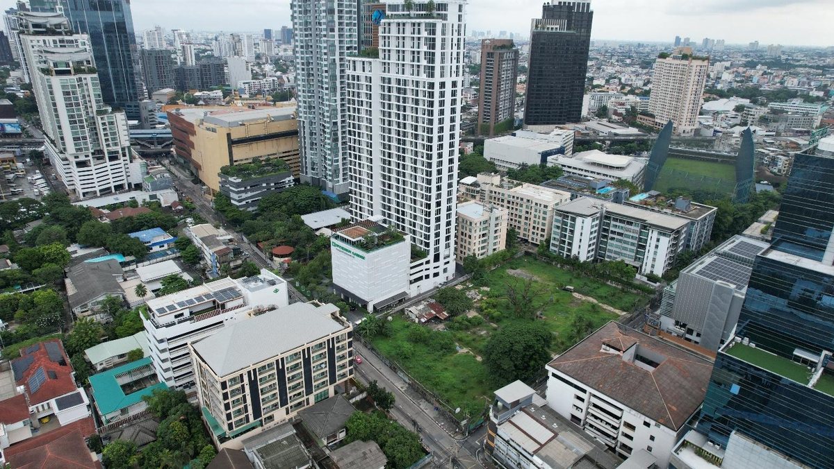 ภาพถ่ายทางอากาศจากโดรนแสดงที่ดินว่างมีต้นไม้และหญ้าปกคลุม ล้อมรอบด้วยอาคารสูงกลางกรุงเทพฯ ก่อนงานสำรวจภูมิประเทศ