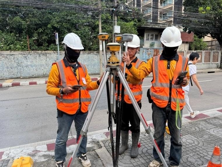 ทีมสำรวจ SPN สามคนตั้งเครื่องรับสัญญาณดาวเทียม GNSS บนขาตั้งริมถนน ตรวจค่าระดับบนหมุดอ้างอิงก่อนเก็บข้อมูล