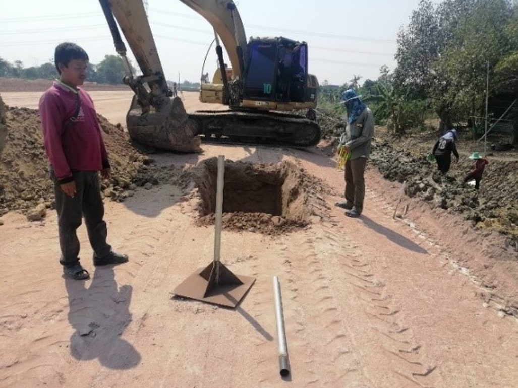 ชุด Settlement Plate แผ่นเหล็กฐานสี่เหลี่ยมเสริมครีบทรงพีระมิดพร้อมท่อวัดตั้งฉาก และท่อเหล็กสำหรับต่อเพิ่มวางอยู่ข้าง ๆ บนพื้นที่งานถมดิน มีรถขุดและหลุมขุดอยู่ด้านหลัง
